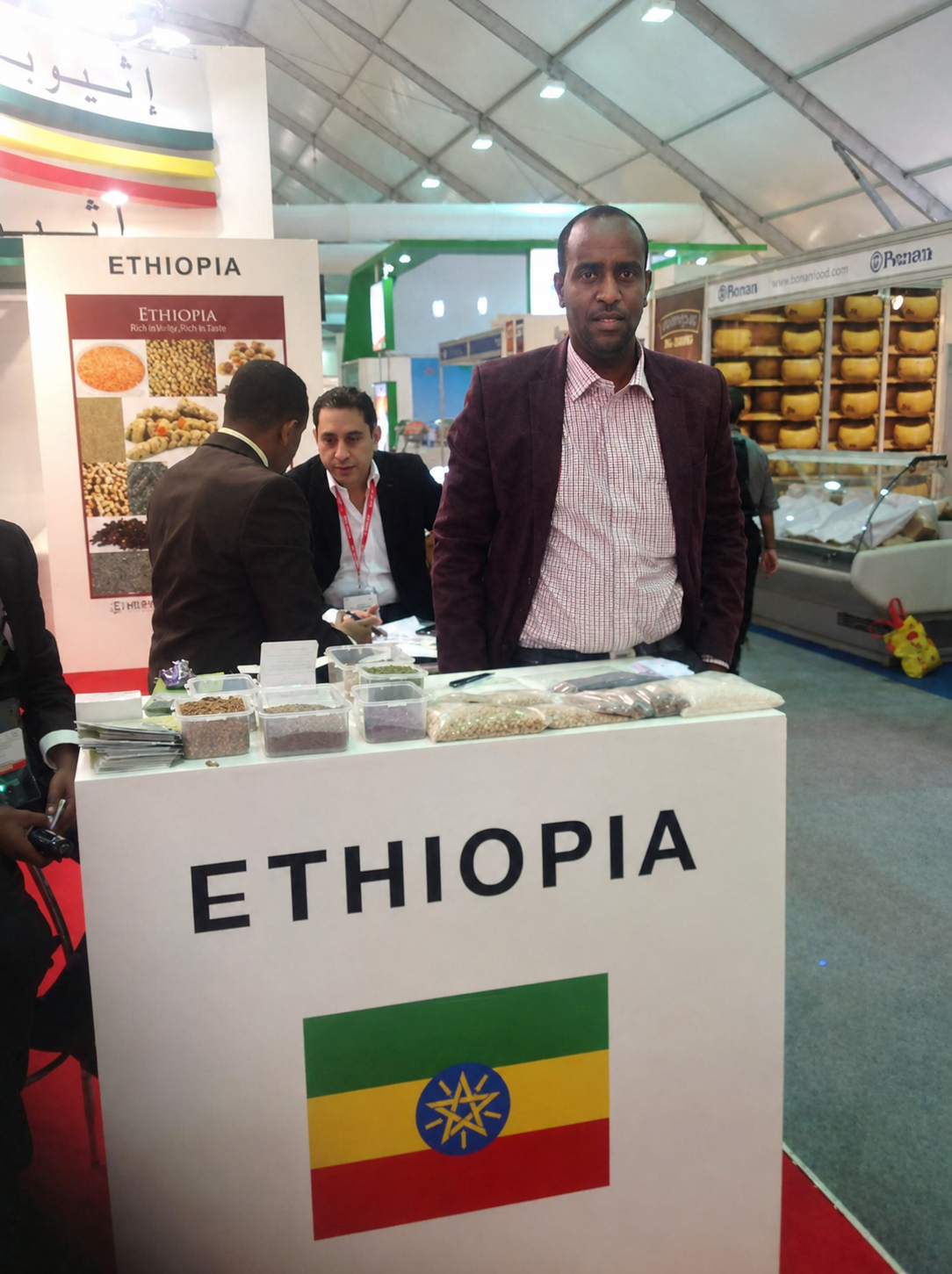 Abdulmuen Mohamed standing behind an exhibition counter marked ETHIOPIA, with Ethiopian origin product samples displayed in front of him.